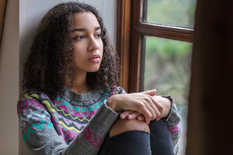 A Black/African American girl sits next to a window and looks off into the distance.