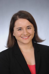 Monica Porter a white woman with brown straight hair, smiles at the camera while wearing a black blazer with a red blouse. The background is a white and grey gradient, as this photo is a headshot.